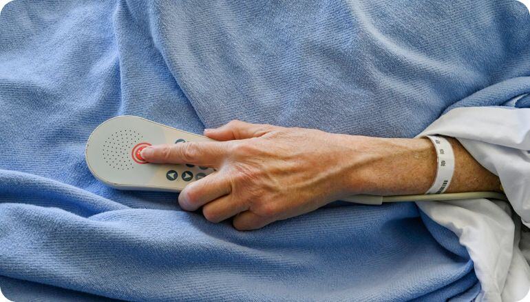 En una cama de hospital, una mano femenina oprime el botón de un dispositivo de llamado a enfermería. 