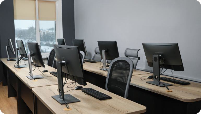 Dos filas de mesas de trabajo con PCs y sillas dentro de una sala que parece pertenecer a un instituto o centro corporativo 