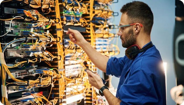 Un técnico con gafas de protección instala elementos de un rack mientras revisa un extenso cableado en un centro de datos. 