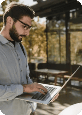 Un colaborador joven de TI con barba y anteojos, toma de pie una lectura desde su laptop en el exterior de su oficina. 